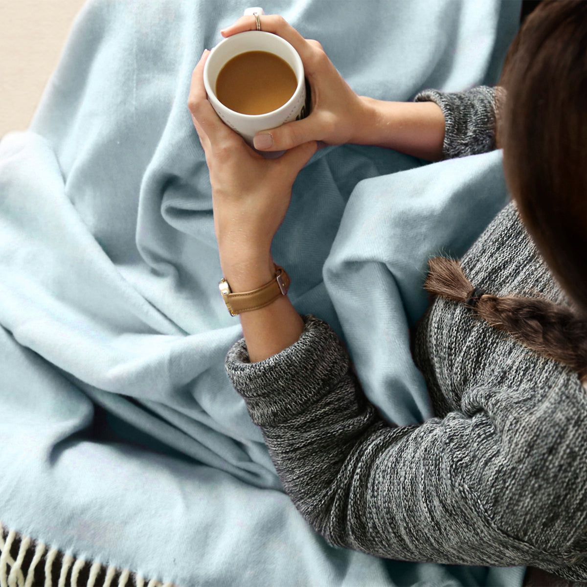 A person wrapped in a soft light blue Italian Herringbone blanket, holding a white mug with coffee.