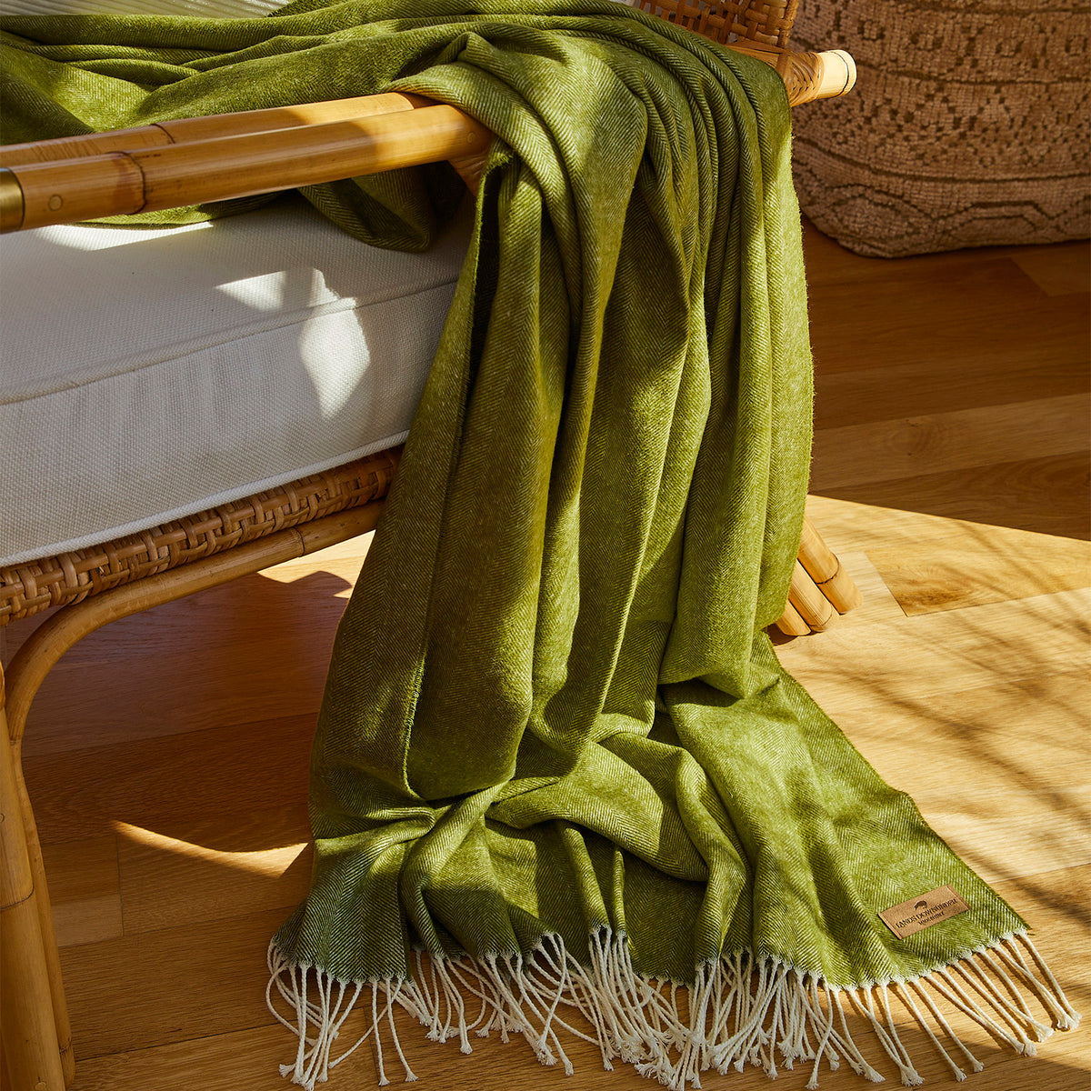 An olive green woven throw blanket with cream fringe is draped over a rattan chair in a sunlit room with a wooden floor.