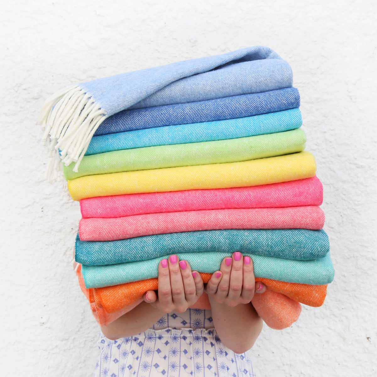 A stack of folded Italian Herringbone throiw blankets in a rainbow of colors, with a light blue fringed blanket on top, held by a woman
