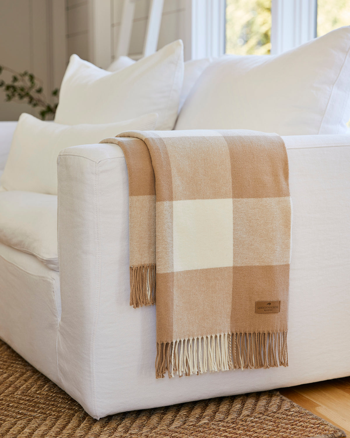 A beige and cream buffalo plaid throw blanket with fringe is draped over the arm of a white upholstered sofa in a bright, modern living room.