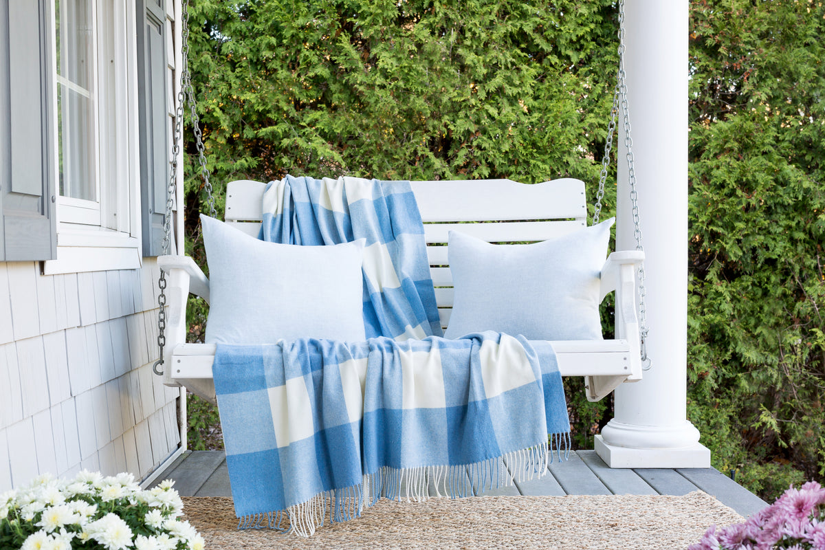 A white wooden porch swing on a porch with a blue and white buffalo plaid throw blanket and matching pillows