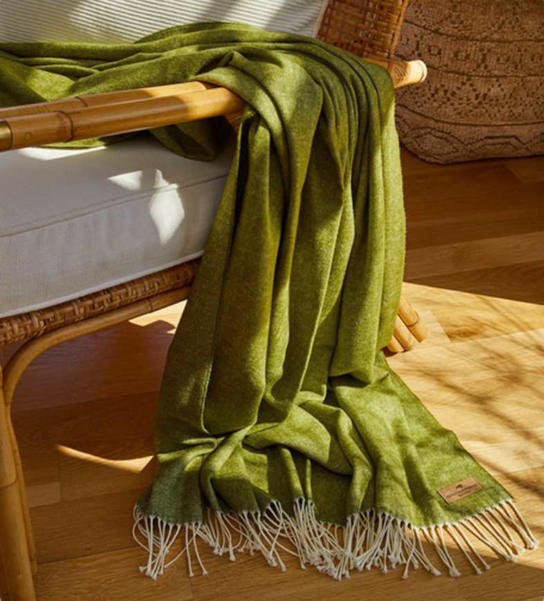 An olive green woven throw blanket with cream fringe is draped over a rattan armchair in a sunlit room with a wooden floor.