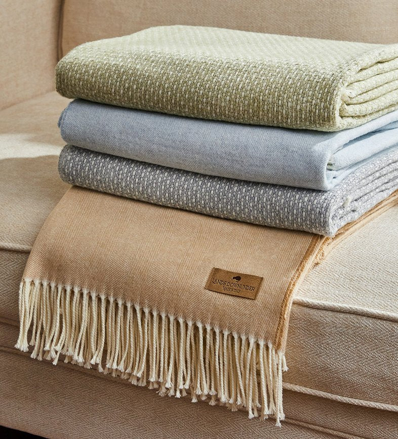 Three folded woven throw blankets in light olive green, pale blue, and beige with cream fringe, stacked on a matching beige sofa. A small tan brand patch is visible on the front blanket.