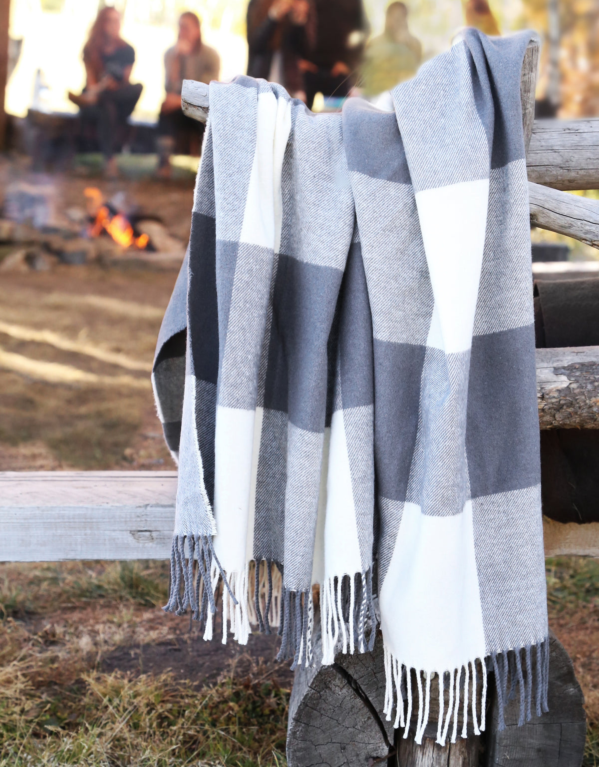A large gray and white buffalo plaid throw blanket with fringe, draped over a rustic wooden fence in an outdoor campfire setting.